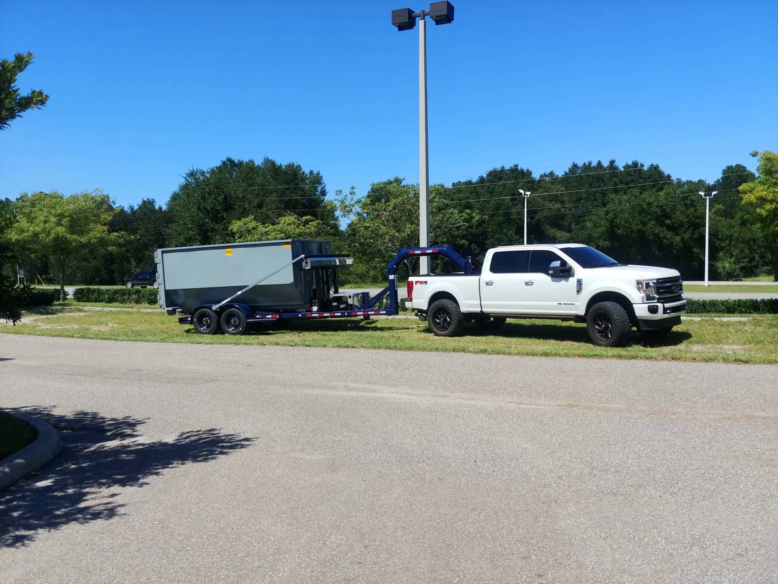 0930221334 1 scaled - Yankee Dumpster LLC White work truck pulling small Yankee dumpster down the road alongside grass easement in Pine Hills, FL